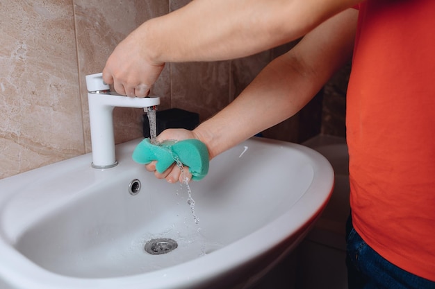 Мужские руки ополаскивают чистящую губку под водой, текущей из крана в раковине, сжимая ее