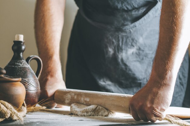 Male hands roll dough and flour with rolling pin next to clay pot and oil bottle on dark table, while cooking