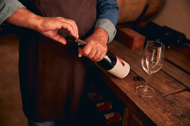 Photo male hands using corkscrew to open bottle of alcoholic drink while man standing by wooden table with empty wineglass