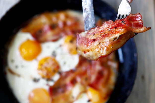 Male hands with a knife and a fork are cutting bacon and eggs in a frying pan.