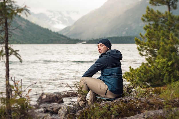 A male hiker and adventurer sits on the shore of a picturesque mountain lake and admires the landscape