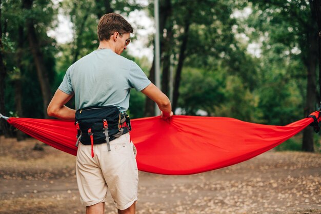 夏のカジュアルな服装の男性ハイカーは森の中でハンモックをまっすぐにします