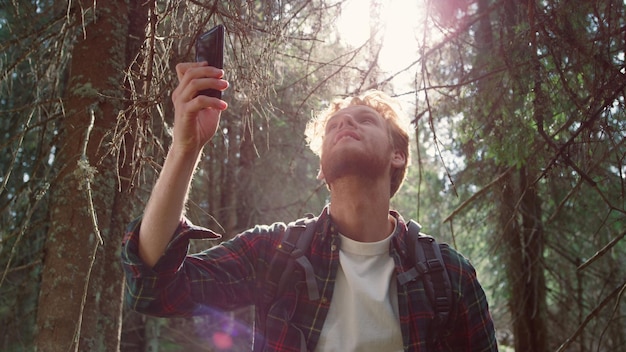 Male hiker with backpack trying to find mobile network in forest Redhead guy holding smartphone in hand Smiling man using mobile phone in woods Handsome tourist standing in green landscape