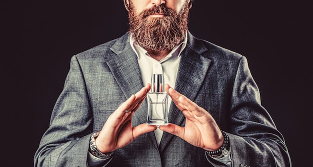 香水のボトルを保持している男性。男の香水、香り。香水またはケルンのボトルと香水、化粧品、香りのケルンのボトル、ケルンを保持している男性。男性的な香水、スーツを着たひげを生やした男