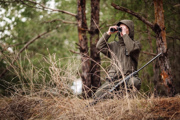 双眼鏡を持った男性ハンターが銃を持って森の中を歩いて狩りをする準備ができています
