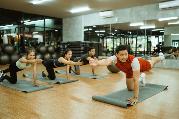 Инструктор-мужчина делает балансирующие движения на коврике с группой спортивных людей в фитнес-зале