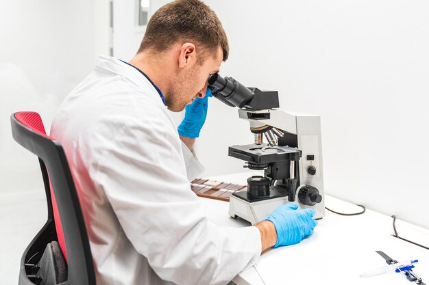 Foto tecnico di laboratorio di sesso maschile in giacca bianca che analizza campioni attraverso un microscopio in un ospedale clinico