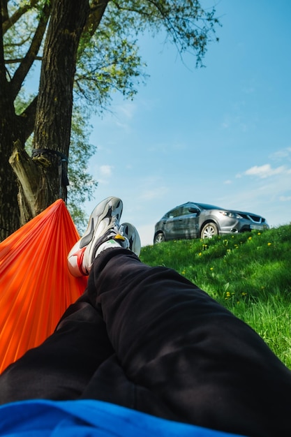 Male legs in a hammock overlooking a parked car on a green lawn in nature