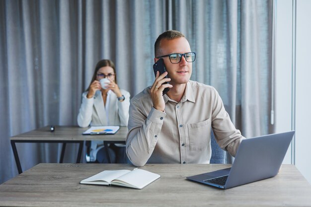 A male manager is talking on a mobile phone while sitting at a table with a computer and a notepad Working atmosphere in an office with large windows