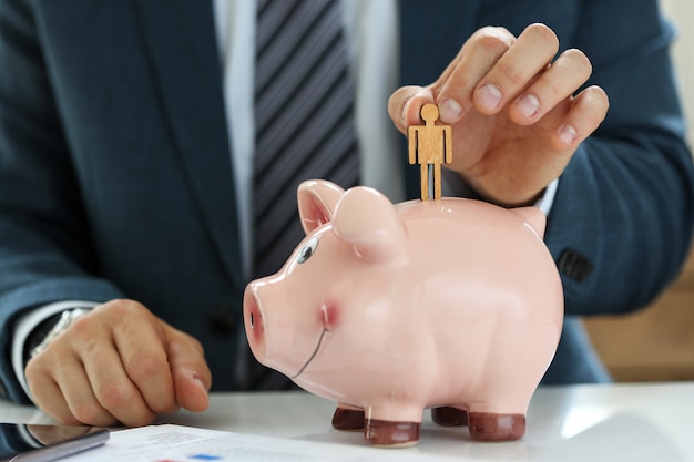 Male manager putting small wooden human figure into piggybank close-up