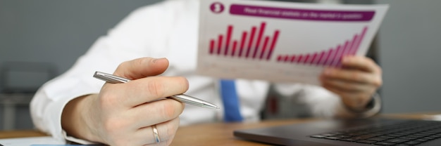 A male manager in a shirt sits at a table and holds a financial report with a diagram