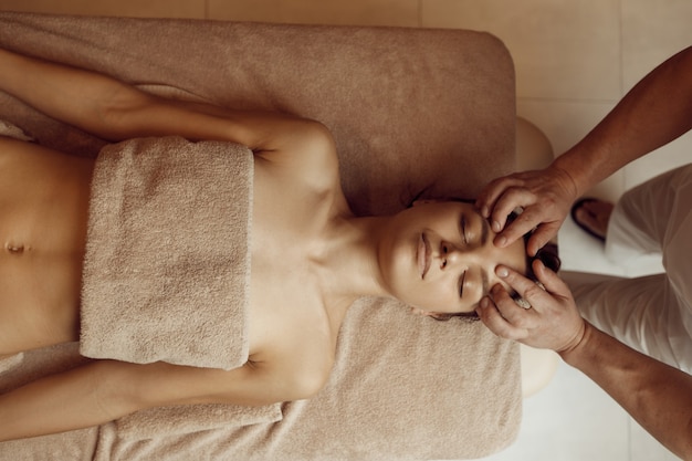 Foto massaggiatore maschio mani sfregamento viso a giovane donna, vista dall'alto, massaggio professionale. massaggio e terapia di rilassamento, cura del corpo e della pelle. signora attraente nel salone della stazione termale