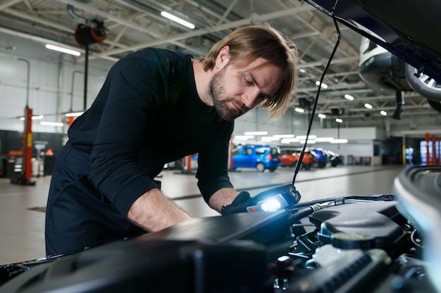 Механик-мужчина в форме с фонариком проводит диагностику под капотом автомобиля Современный и технологичный автосервис