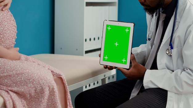 Male medic working with greenscreen on tablet at examination, consulting pregnant person. Doctor using chroma key display with isolated mockup template and blank copyspace. Close up.