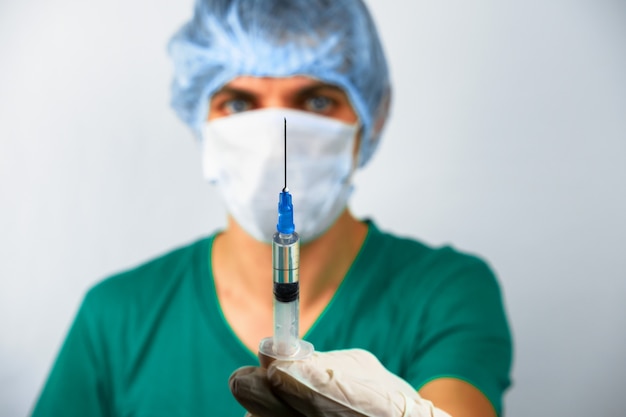 Male medical worker in a cap, mask and gloves holding a syringe