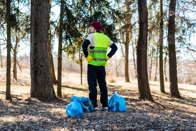 Male nature activist in yellow vest and gloves stands near blue\
trash bags vertical back photo