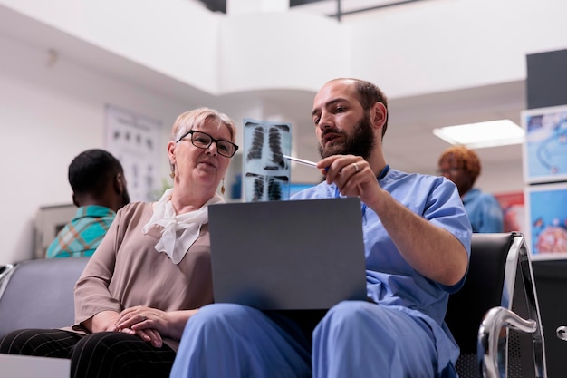 Male nurse explaining x ray diagnosis to old woman in hospital reception lobby, talking about radiography scan results and disease treatment. People chatting about healthcare in waiting area.