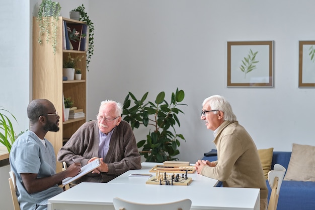 male nurse giving recommendations to senior men while they sitting at the table during chess game in nursing home