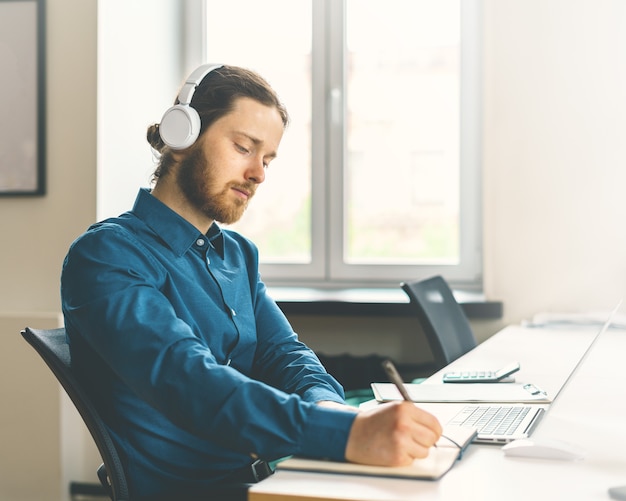 Male office worker dressed in casual clothes in headphones watching online webinar on laptop