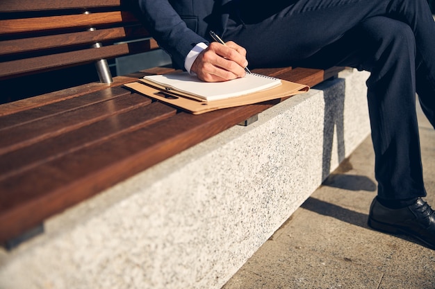 Мужчина в официальной одежде проводит время за работой на скамейке и пишет ручкой