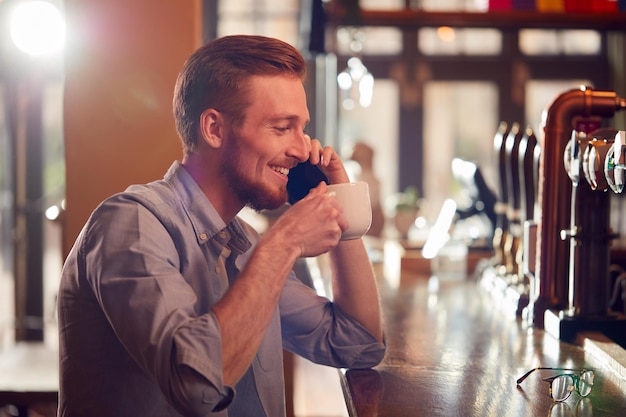 カウンターに座ってコーヒーを飲み、携帯電話で話しているバーの男性の所有者またはマネージャー