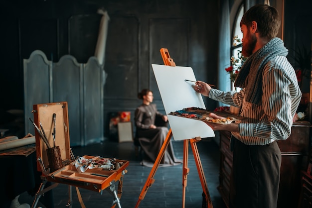 Male painter drawing portrait against female poseur in studio.