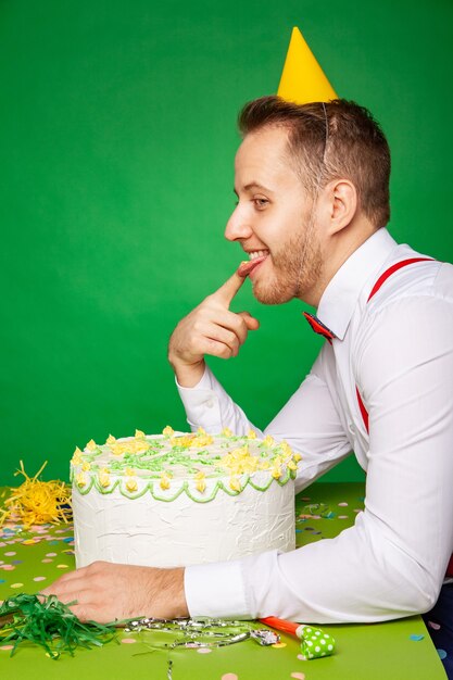 파티 모자를 쓴 남성은 테이블에 앉아 스튜디오의 녹색 배경을 올려다보며 달콤하고 맛있는 생일 케이크를 시식합니다.
