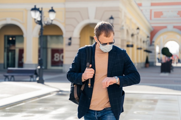 Male passenger checks time on watch waits for bus arrival carries backpack wears protective disposable medical mask protects from Covid19 viruses afraids of infection risk travels alone