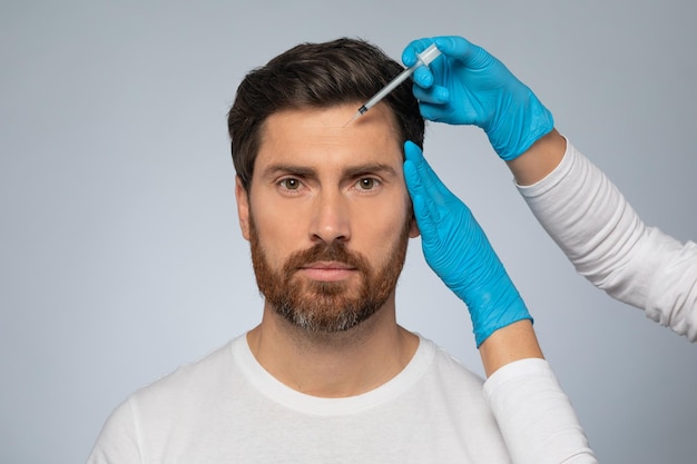Мужчина-пациент косметолога, получающий инъекцию наполнителя в зону лба, хочет выглядеть моложе, заботясь о коже