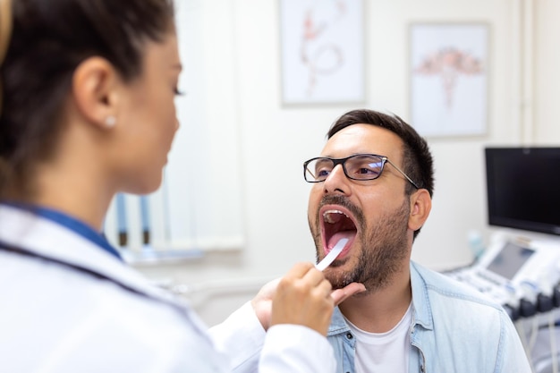 Male patient opening his mouth for the doctor to look at his throat Female doctor examining sore throat of patient in clinic Otolaryngologist examines sore throat of patient