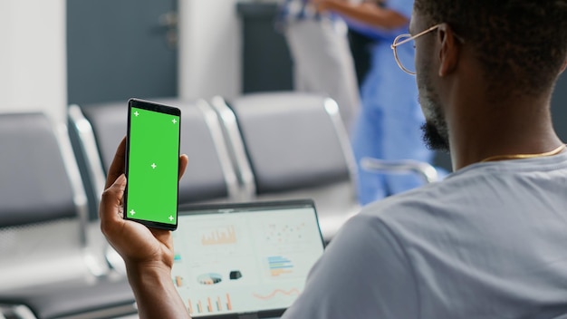 Male patient using greenscreen template on smartphone while he
waits in facility waiting area. young man looking at isolated
mockup display and chroma key, blank copyspace on mobile phone
screen.