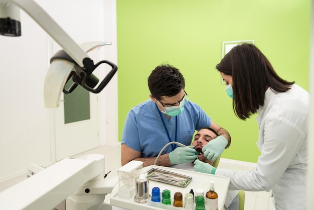 Male Patient With Dentist And Assistant In A Dental Treatment  Wearing Masks And Gloves