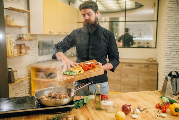 Male person puts vegetables into the frying pan with roasted meat on the kitchen.