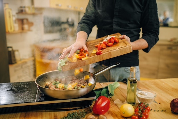 Male person puts vegetables into the pan with meat