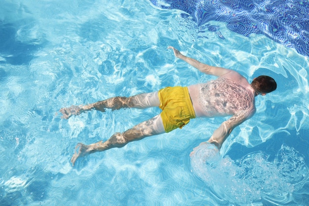 Male person in swimming pants floating on water on belly clear water in pool