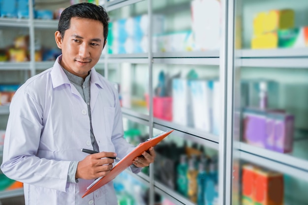 Male pharmacist in uniform writing with pen while holding clipboard standing next to display case