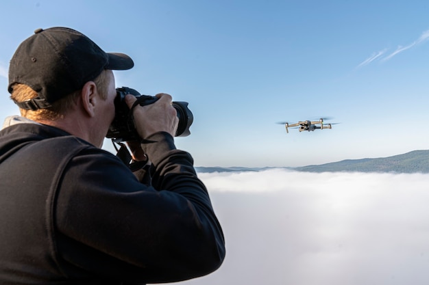 A male photographer takes pictures of a drone hovering in a thick fog in the air above the top of a ...