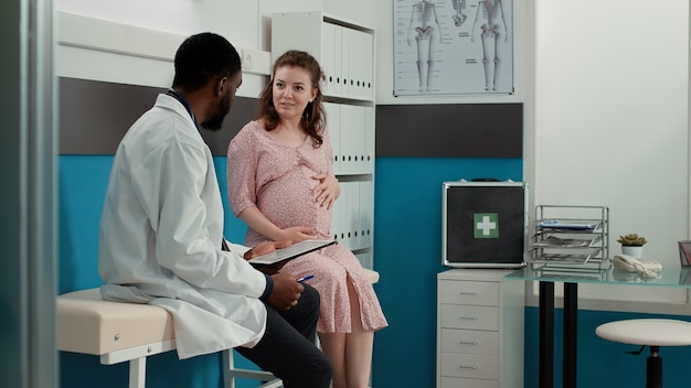 Photo male physician taking notes on checkup files at consultation with expectant patient, having conversation in medical office. health specialist meeting with future mother at appointment.