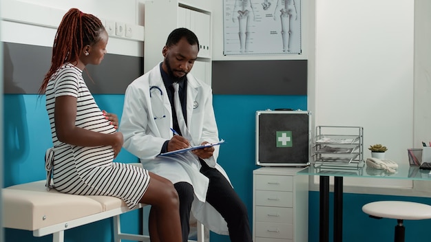 Male physician taking notes at checkup visit with pregnant woman, having private conversation about pregnancy. Medic using health care service documents to give maternity advice in cabinet.