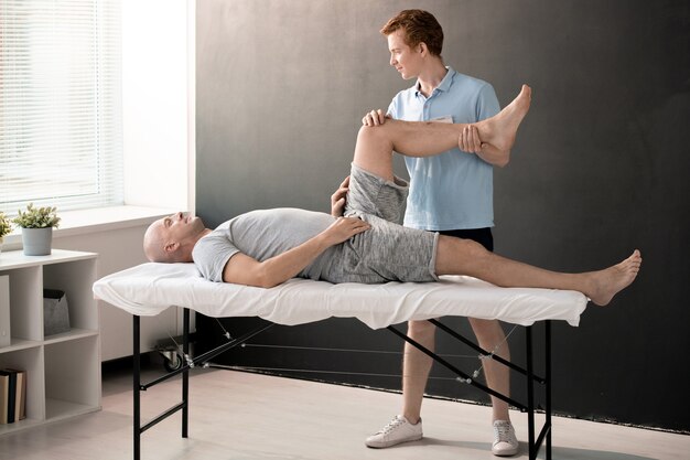 Мужской физиотерапевт держит пациента ногу согнутой в колене, помогая ему с одним из физических упражнений в реабилитационном центре или клиниках