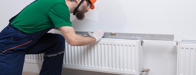 A male plumber installs a radiator in the heating system of an apartment Guy in overalls and a gas wrench