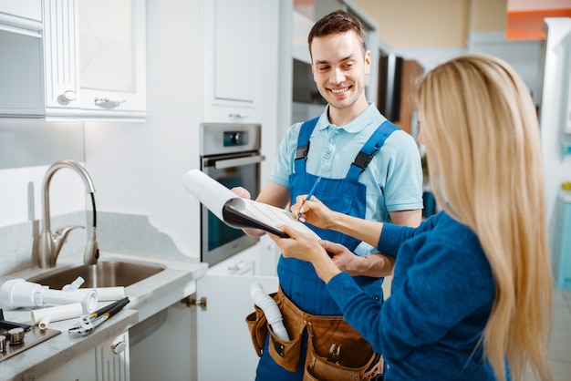 Male plumber in uniform and female customer in the kitchen. Handyman with toolbag repair sink, sanitary equipment service at home