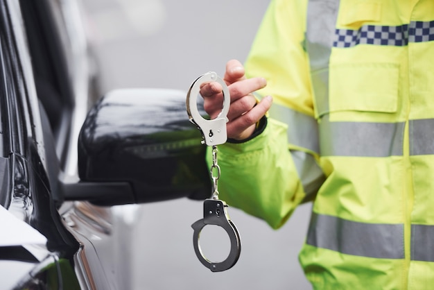 Male police officer in green uniformstanding with handcuffs near automobile.