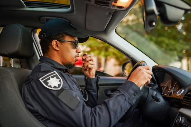 写真 パトカーでウォーキートーキーで話している制服とサングラスの男性警官。警官は法律を守ります。警官は街の通り、秩序と正義の管理に取り組んでいます