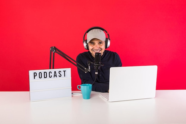 Male presenter with cap in podcast radio recording studio
smiling, next to a computer, a microphone and a light box with the
word podcast, on red background. podcasting, broadcast concept