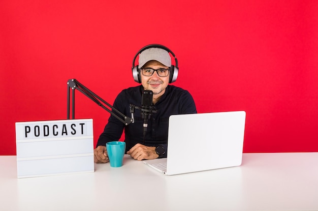 Male presenter with cap in podcast radio recording studio\
smiling, next to a computer, a microphone and a light box with the\
word podcast, on red background. podcasting, broadcast concept