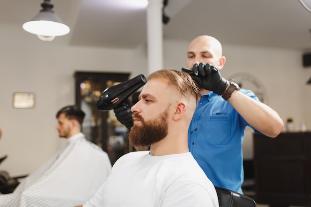 Male professional hairdresser serving client, drying hair with hairdryer