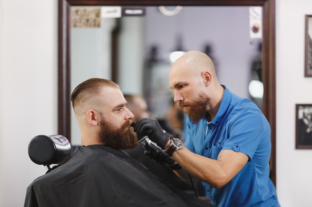 Parrucchiere professionista maschio che serve cliente, rasatura barba folta e grande rasoio a mano libera