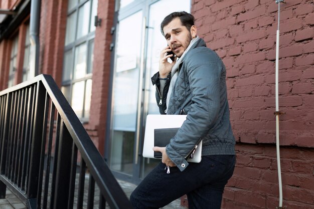 A male programmer talking on the phone with a laptop in his hands enters the office building