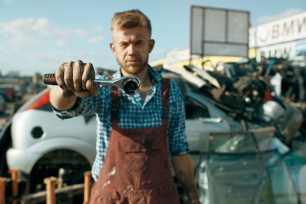 Мужчина-ремонтник показывает гаечный ключ на свалке автомобилей. Автомобильный лом, автомобильный утиль, автомобильный мусор. Брошенный, поврежденный и раздавленный транспорт, свалка
